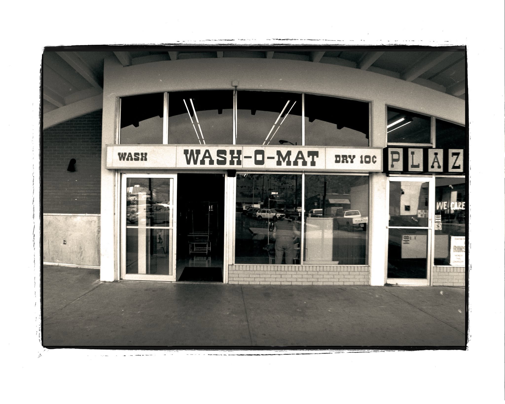 Black-and-white photo of a vintage “WASH-O-MAT” laundromat with glass doors and storefront windows; signs above read “Wash” and “Dry 10¢.” Reflections of cars and the street are visible.