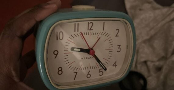 A hand holds a small blue analog alarm clock showing the time as 6:44:37. A beige patterned cloth and a black object are in the background.