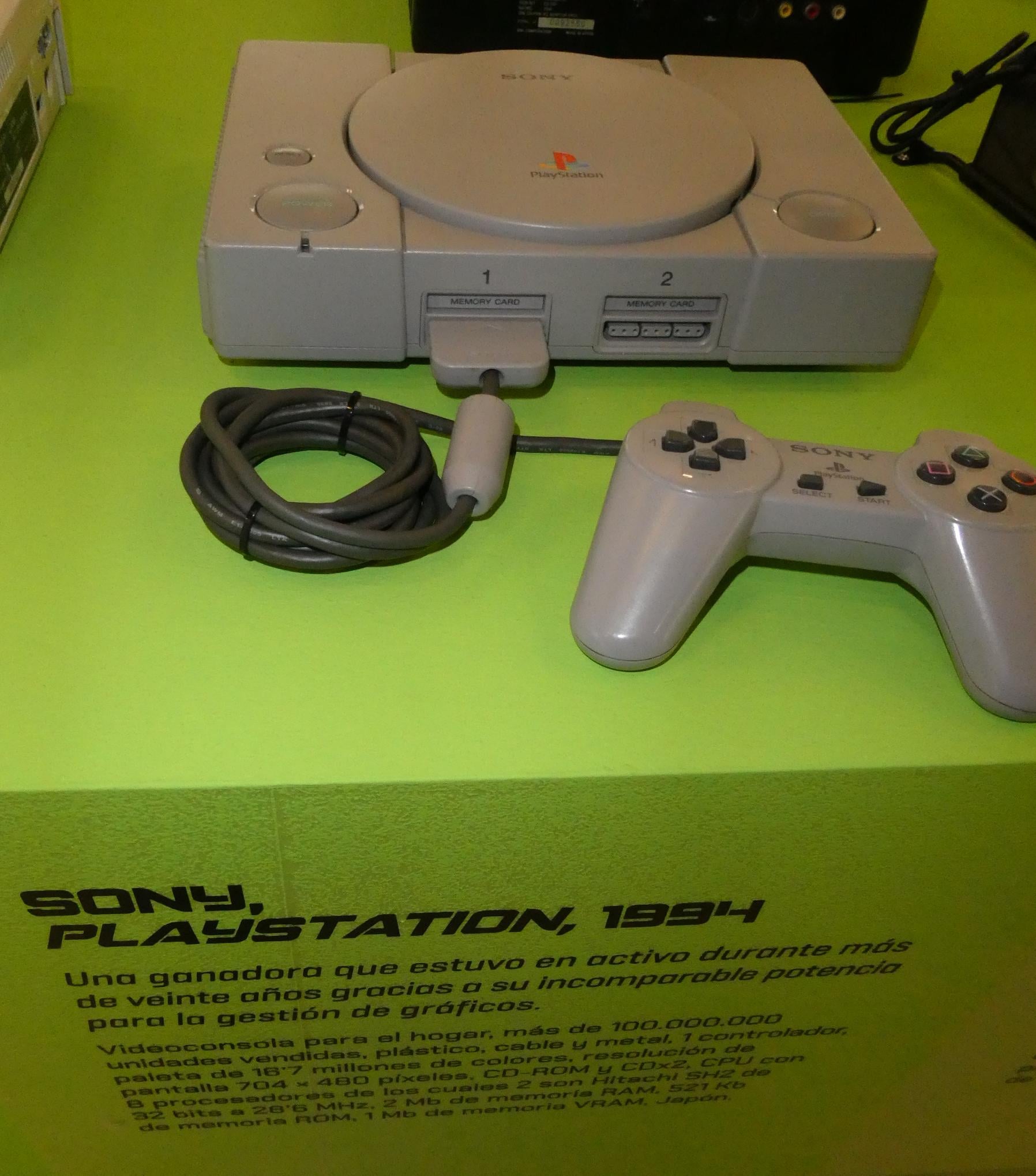A gray Sony PlayStation console from 1994 with a matching controller is displayed on a green surface with a description about its release and features in Spanish.