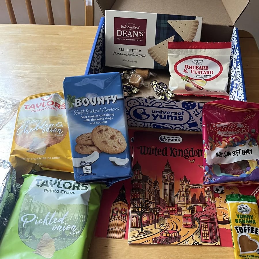 A selection of British snacks, including potato crisps, cookies, candies, and shortbread, displayed around a Universal Yums UK box and illustrated pamphlet on a wooden table.