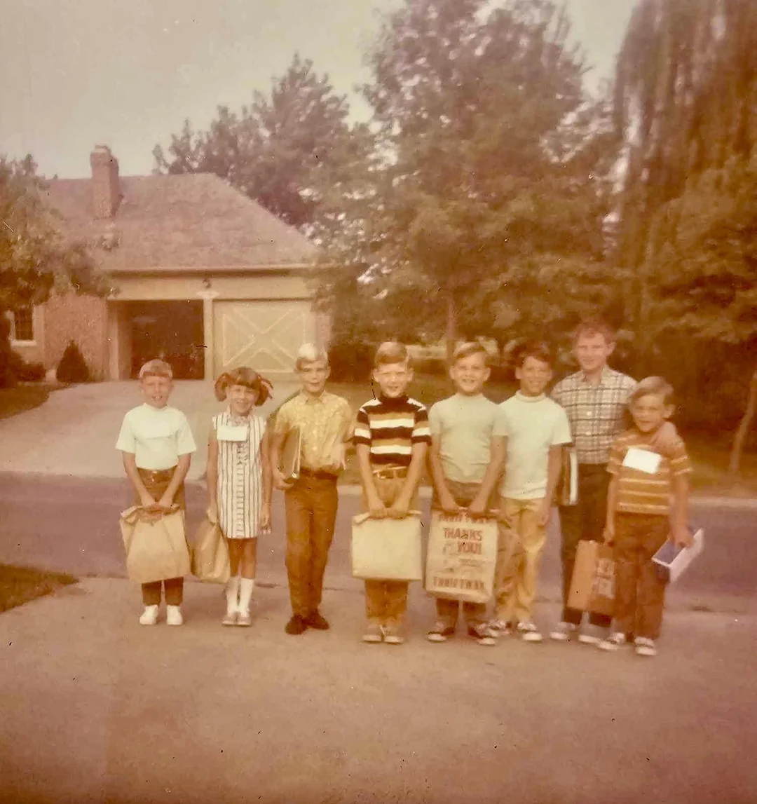 Eight children stand in a row on a driveway holding paper bags. A house with a garage, trees, and a lawn are in the background. The image has a faded, vintage look.