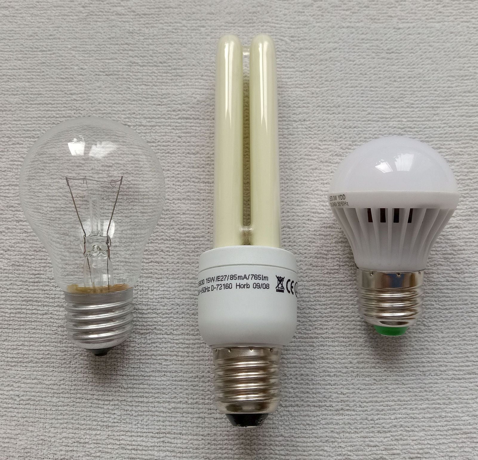 Three light bulbs on a white textured background: an incandescent bulb on the left, a compact fluorescent bulb in the center, and an LED bulb on the right.