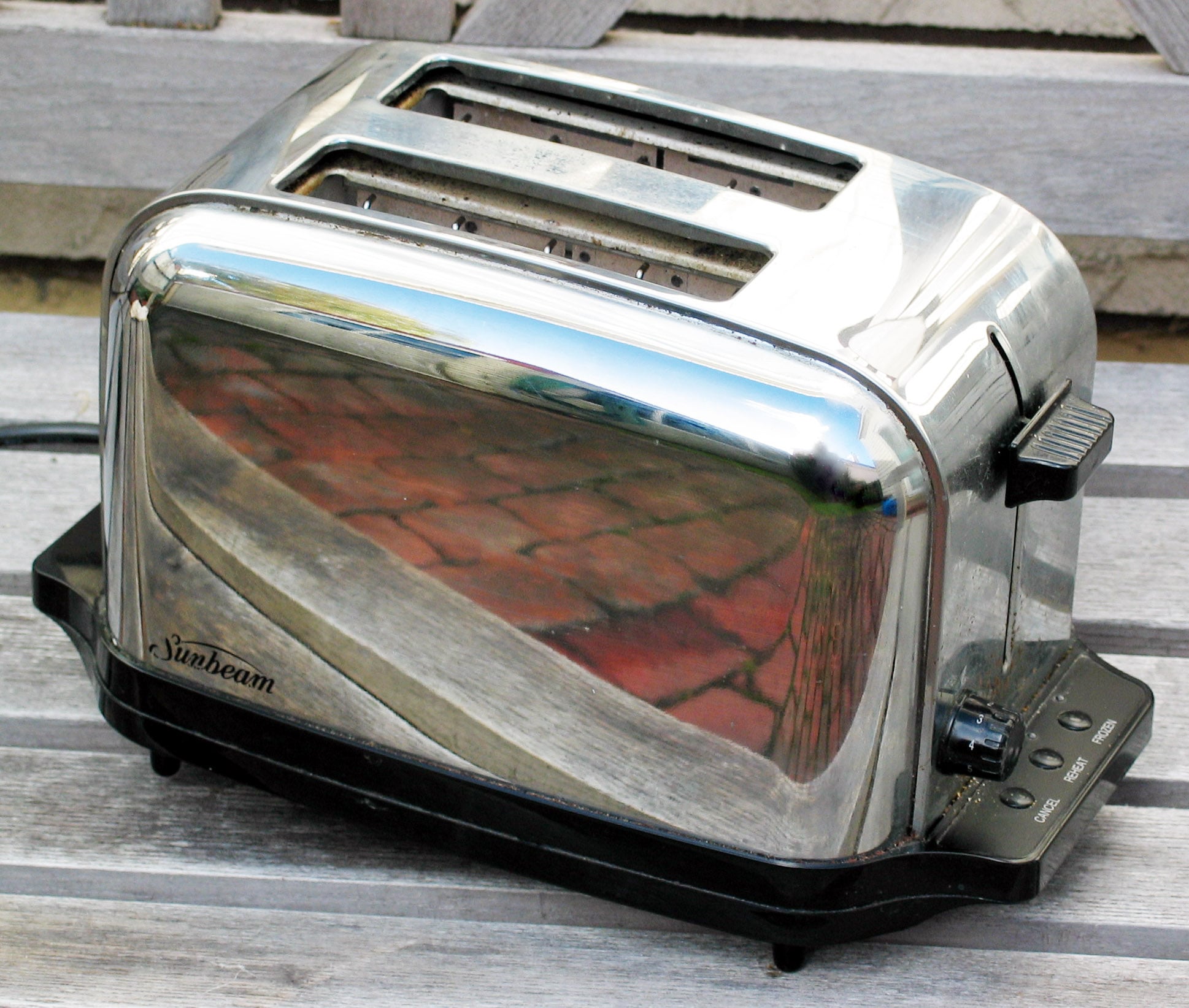 A shiny stainless steel Sunbeam toaster with two slots sits on a wooden surface, reflecting the surroundings. The controls and lever are visible on the side.