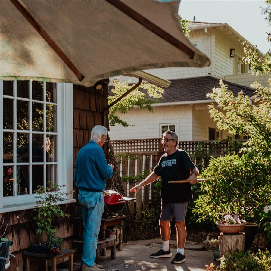 Two older men stand on a sunny patio near a small grill, chatting. One holds grilling tools, the other faces him. Green plants and a wooden fence surround them, with houses visible in the background.
