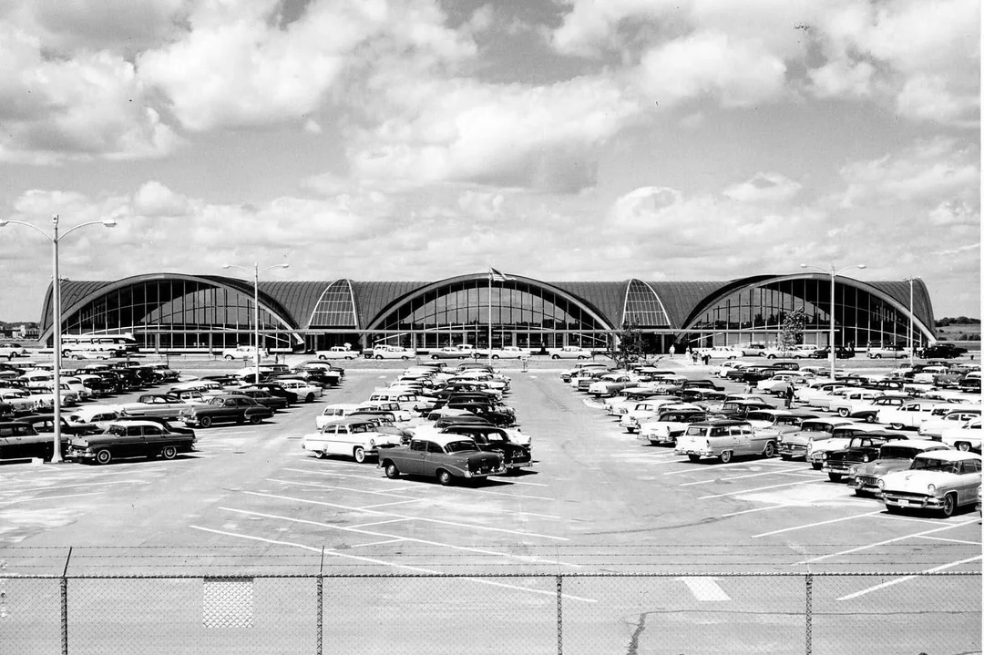 Black and white photo of a mid-century airport terminal with arched roofs and large glass windows, seen from a parking lot filled with classic 1950s cars under a partly cloudy sky.