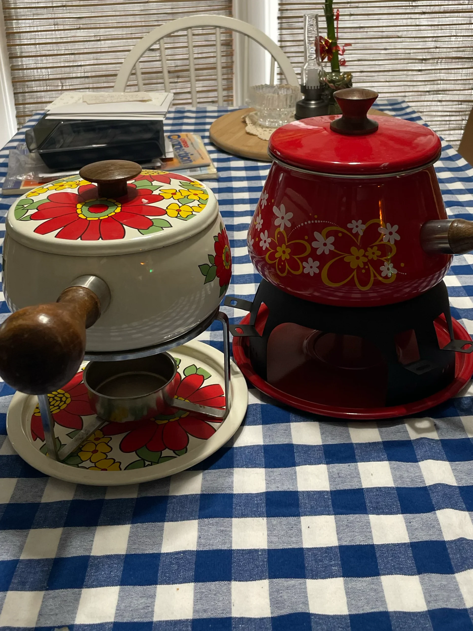 Two vintage fondue pots with floral designs sit on a blue and white checkered tablecloth; one pot is red with yellow flowers, the other is white with red flowers.