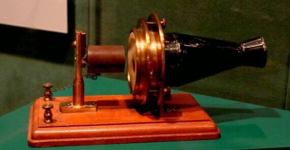 An antique microphone (carbon transmitter) with a brass and black cone, mounted on a wooden base with metal components, displayed against a green background.