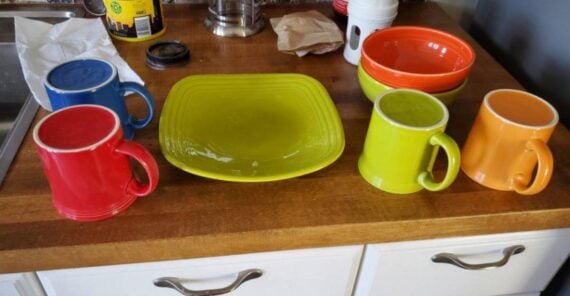 Four colorful mugs, three bowls, and a square plate sit on a wooden countertop in front of white drawers. There’s a coffee press, paper towel, and canisters in the background.