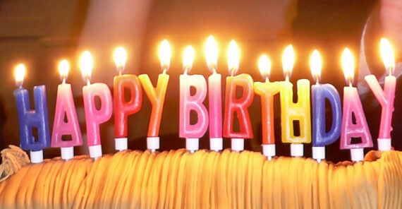 Colorful candles spelling “Happy Birthday” are lit atop a cake with frosting. Two people in dark clothing stand in the background, partially visible.