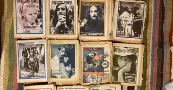 Ten vintage Rolling Stone magazines with illustrated and black-and-white covers are arranged on a wooden surface. Each cover features portraits or artistic designs. The magazines appear aged and worn.