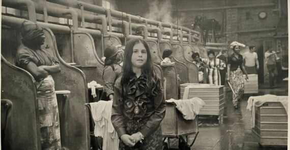 A young girl stands in the foreground of an old laundry facility, with women working at washing stations in the background. The scene appears busy and industrial, with pipes, steam, and baskets visible throughout.