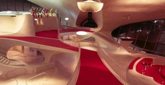 Spacious, modern interior with sweeping red carpet, curved white railings, flag display, chandeliers, and large windows letting in natural light.
