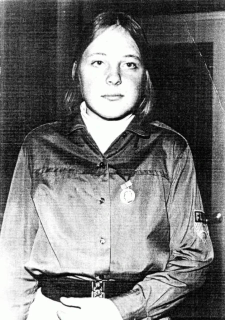 Black and white photo of a young woman with shoulder-length hair, wearing a buttoned shirt with a medal and a patch on the sleeve, standing indoors and looking at the camera.