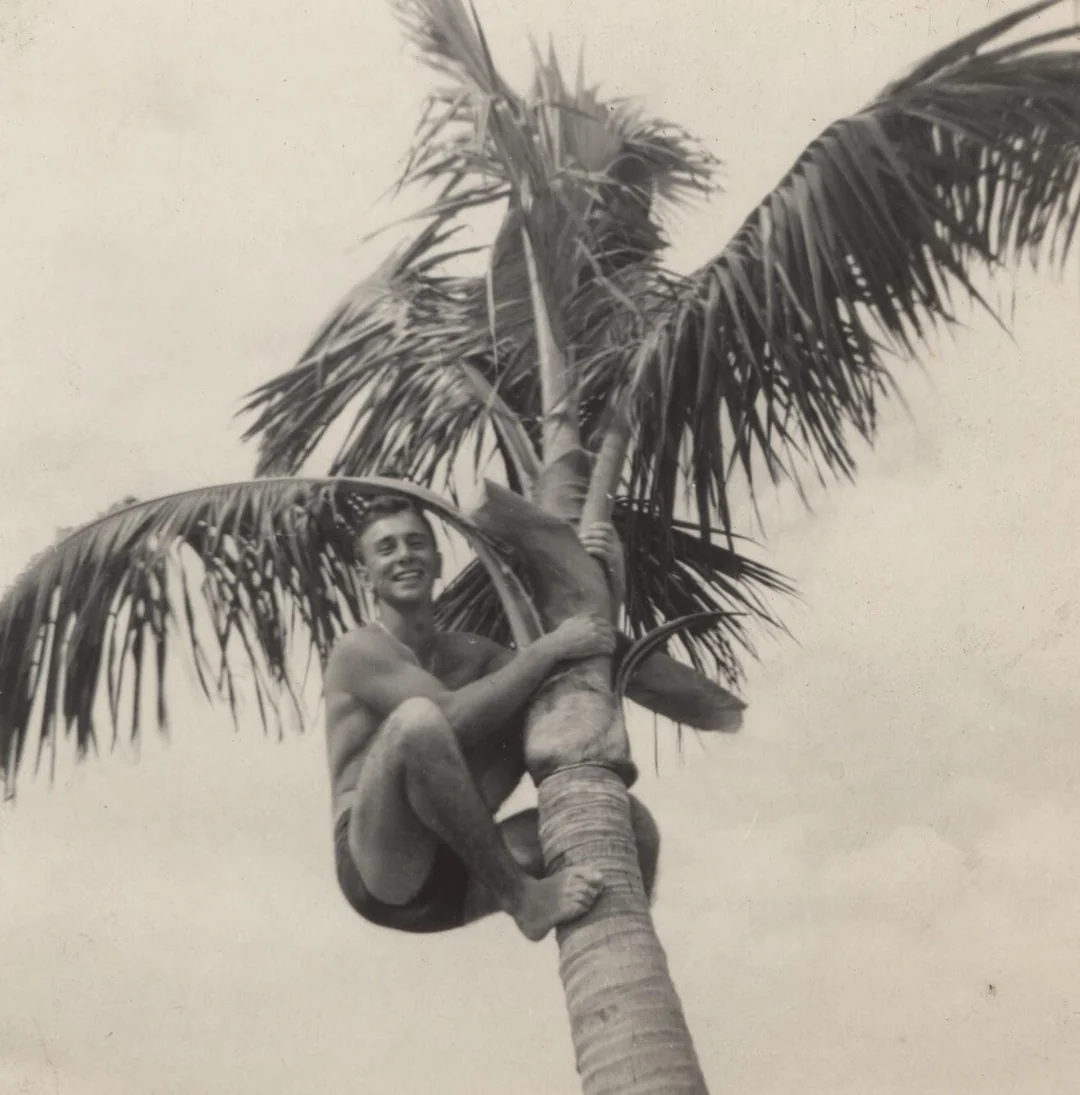 A man in swim trunks smiles while climbing a palm tree. The sky is clear and bright, giving a tropical, playful atmosphere. The photo is black and white.