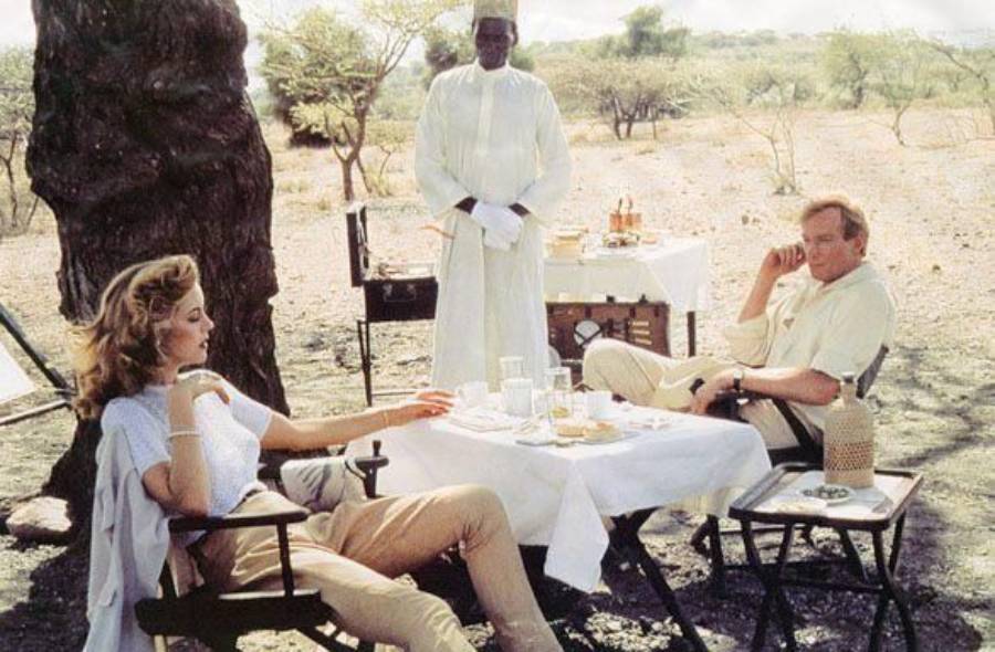 A woman and a man sit at a white-clothed outdoor table with drinks and food, while a person in white robes stands nearby. The setting is sunny, dry, and surrounded by sparse trees.
