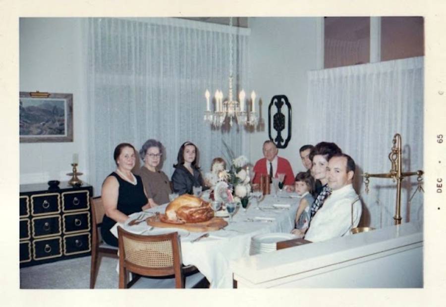 Eight people sit around a dining table set for a meal with a large roasted turkey as the centerpiece. The room is decorated with a chandelier and sheer curtains. The photo is dated December 1965.