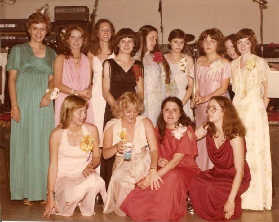 A group of thirteen young women in formal dresses pose together indoors, some standing and some kneeling, with musical instruments visible in the background.