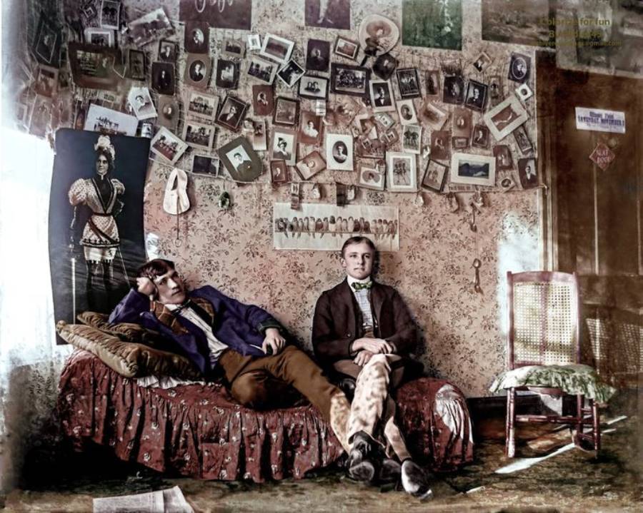 Two young men sit on a patterned sofa in a vintage room. The wall behind them is covered with many photos and portraits. A rocking chair and a tall photo of a person in costume are also visible.