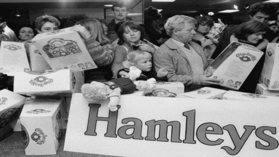 Shoppers crowd around a display of Cabbage Patch Kid dolls at Hamleys toy store, holding boxes and looking eager. A child sits among the crowd, and several dolls and boxes are visible.