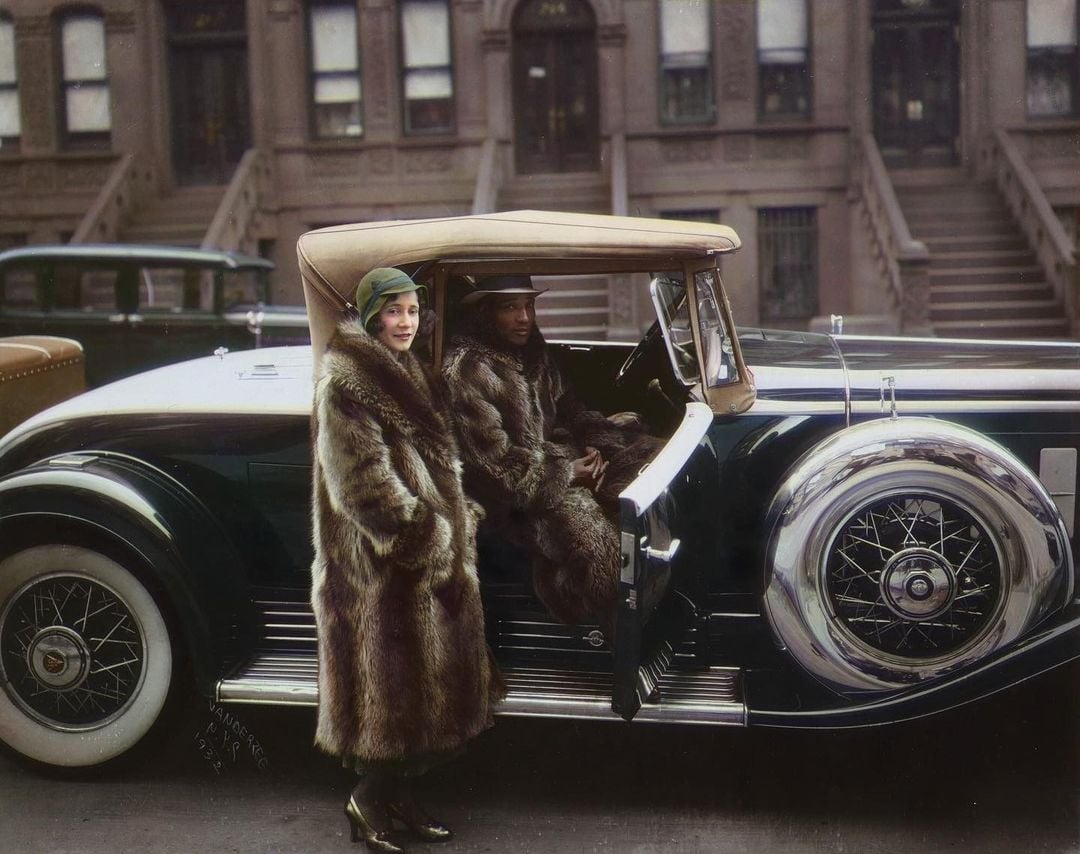 A woman in a fur coat stands by a vintage car, while a man, also wearing a fur coat, sits inside. They are parked on a city street in front of old brownstone buildings.