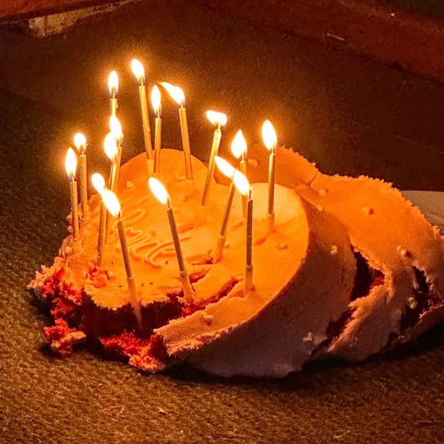 A heart-shaped birthday cake with lit candles has fallen and is smashed on the floor, but the candles are still burning.