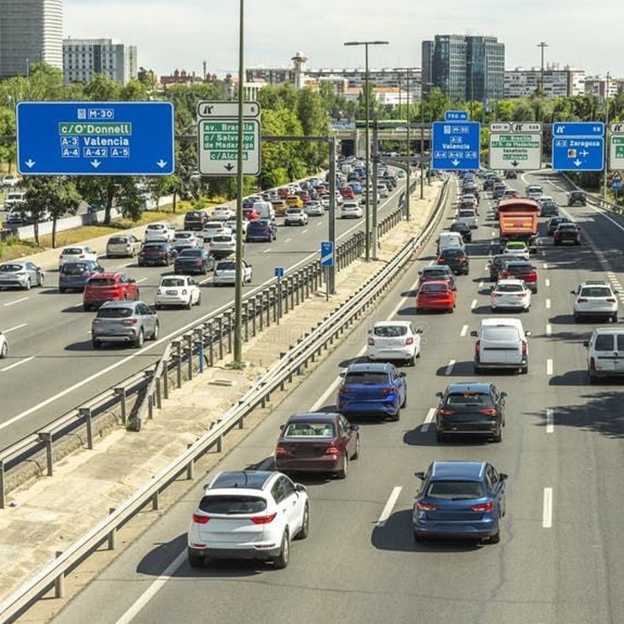 A busy multi-lane highway with heavy traffic, road signs in Spanish, cars traveling in both directions, and city buildings in the background under a clear sky.