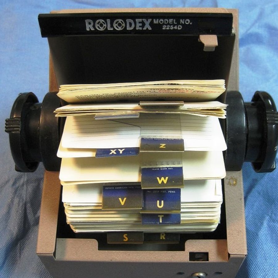 A classic Rolodex card file with alphabetical tabs and handwritten cards, used for organizing contact information, sits open on a blue surface.
