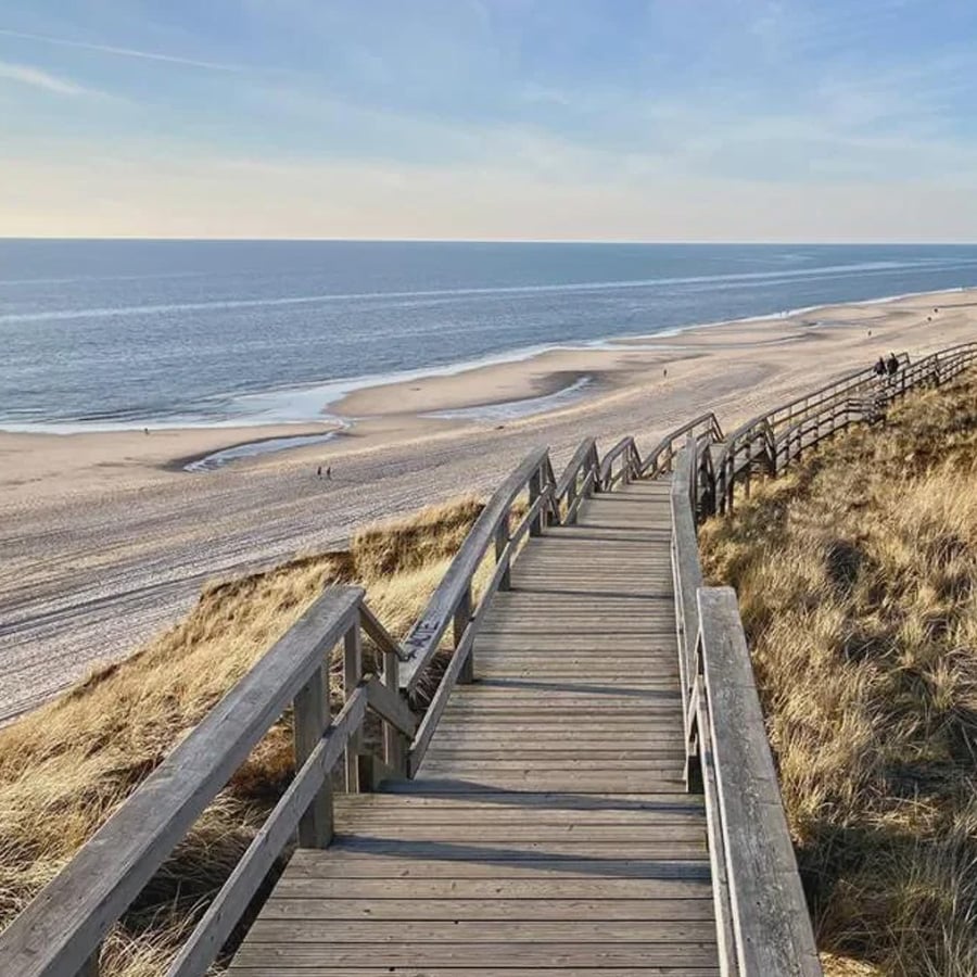A wooden boardwalk with railings leads down through grassy dunes to a sandy beach, with the calm sea and blue sky in the background. A few people are visible walking along the shore.