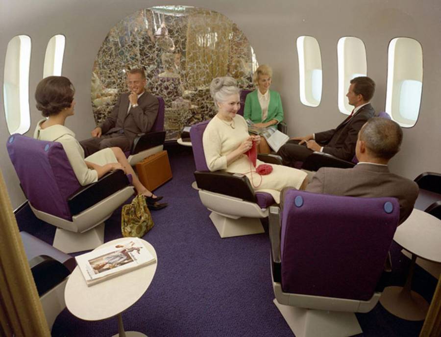 Six people in formal attire sit and talk in a spacious, retro airplane lounge with blue carpet and purple chairs. A woman is knitting, and magazines are on a nearby table. The setting appears to be from the 1960s or 1970s.