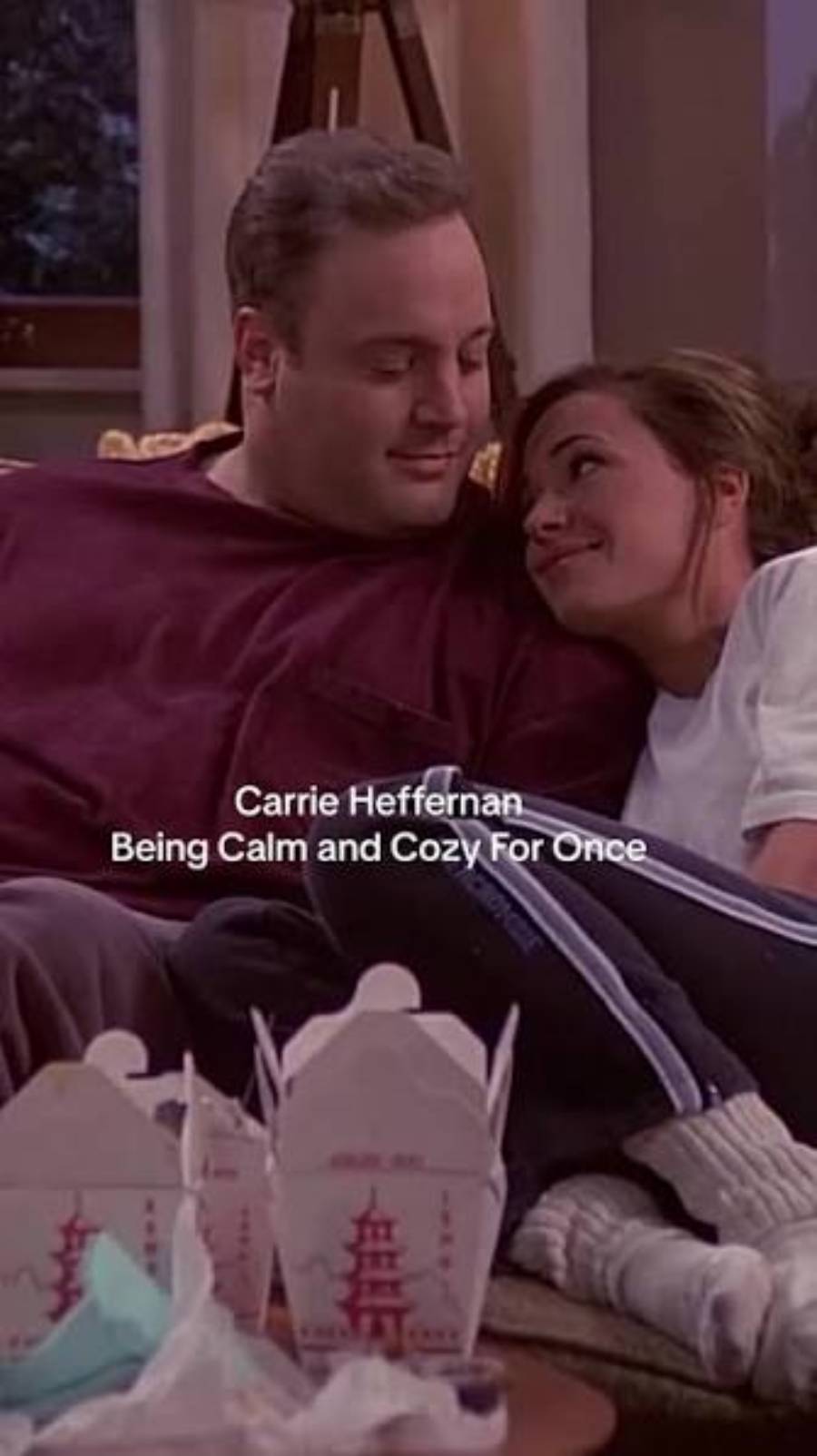 A man and woman relax together on a couch, smiling at each other. Chinese takeout boxes sit on the table in front of them. Text on the image reads: “Carrie Heffernan Being Calm and Cozy For Once.”