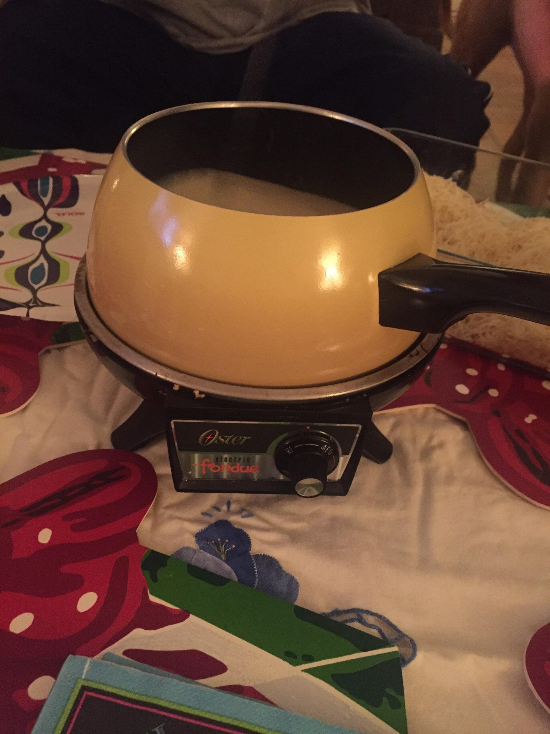 A yellow electric fondue pot filled with melted cheese sits on a colorful tablecloth, with the control knob on the base visible in front.