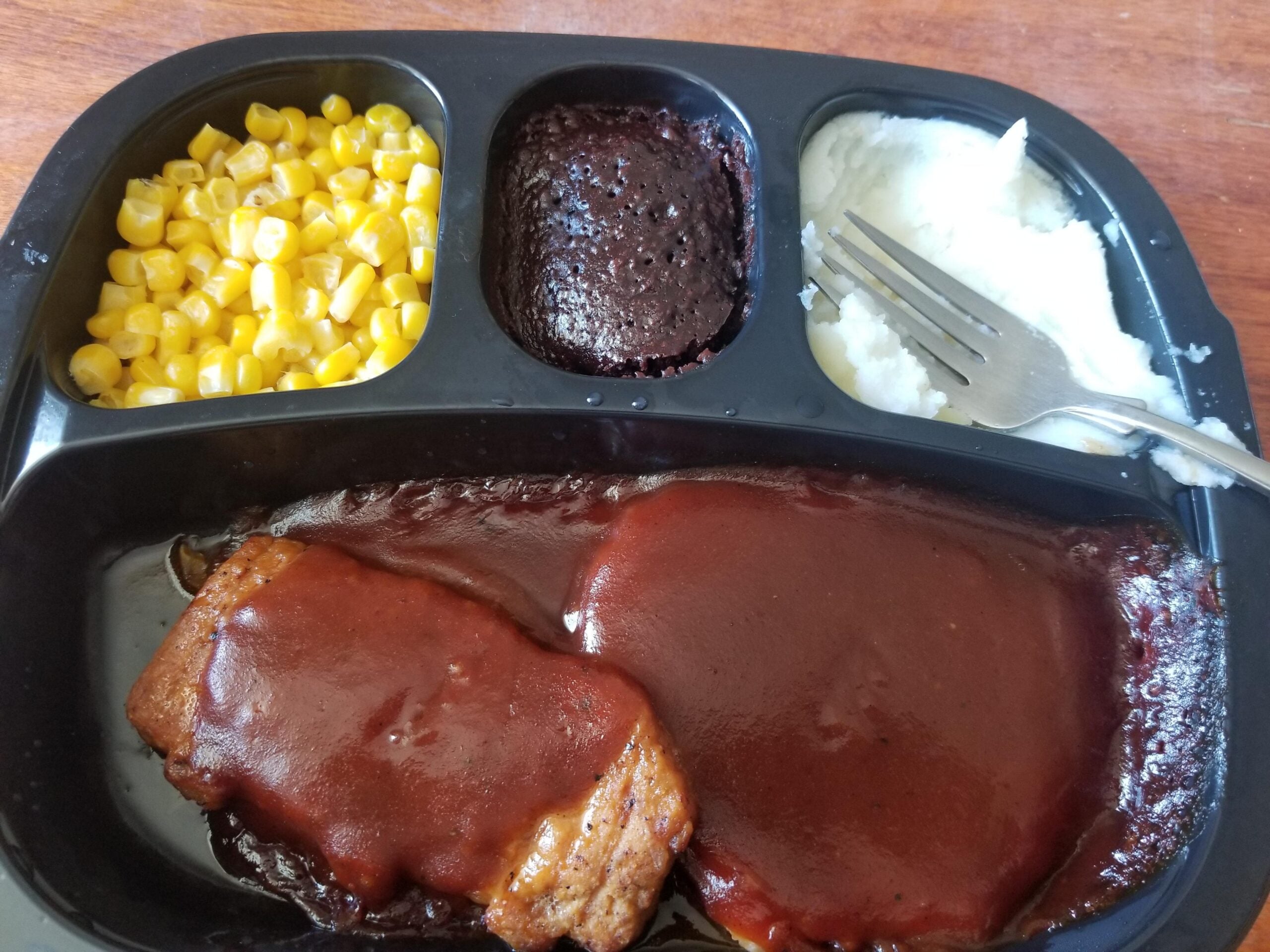 A TV dinner with corn, a brownie, mashed potatoes with a fork, and a piece of meatloaf covered in brown sauce, all in separate compartments on a black tray.