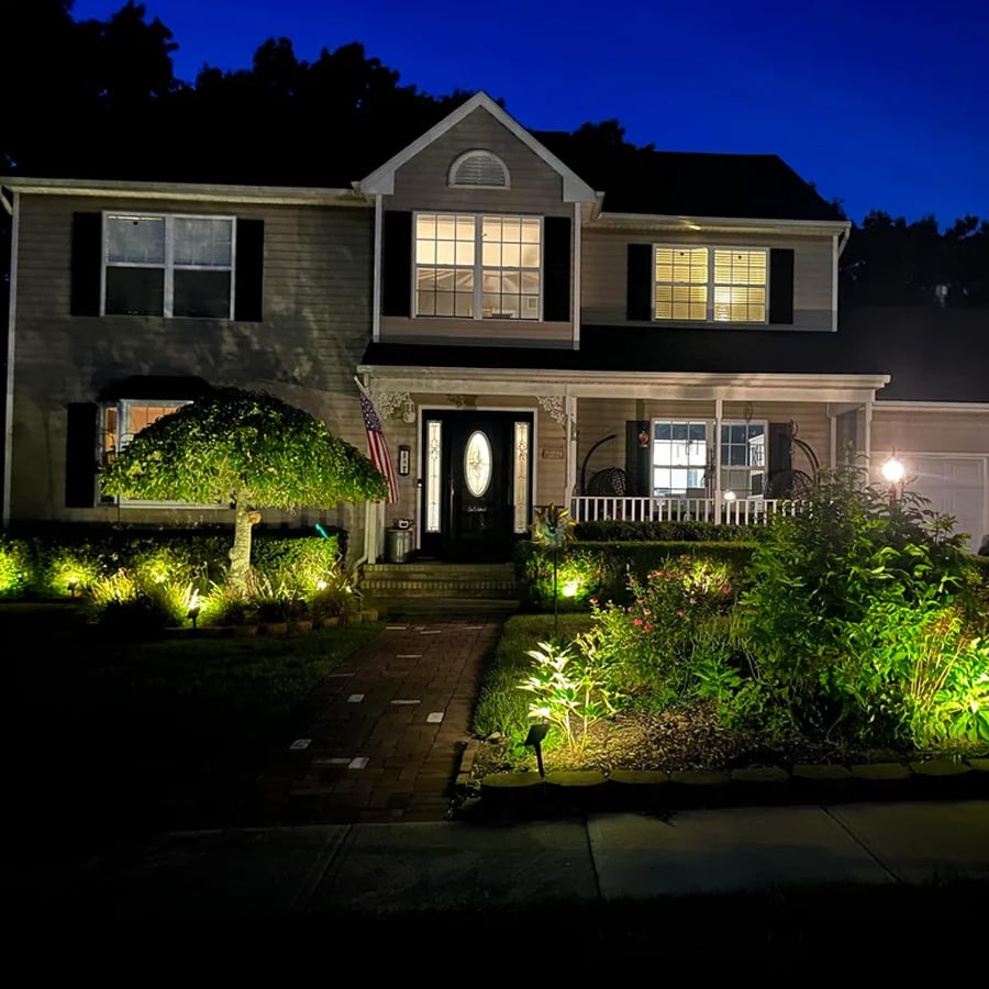 A two-story house at night with lights illuminating the front yard, garden, and pathway. Several windows are lit, and a tree and bushes line the walkway to the front door.