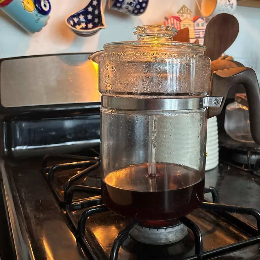 Glass stovetop coffee percolator with dark coffee brewing, sitting on a gas burner. Steam is visible inside the lid. Colorful cups hang above in the background, kitchen utensils on the right.
