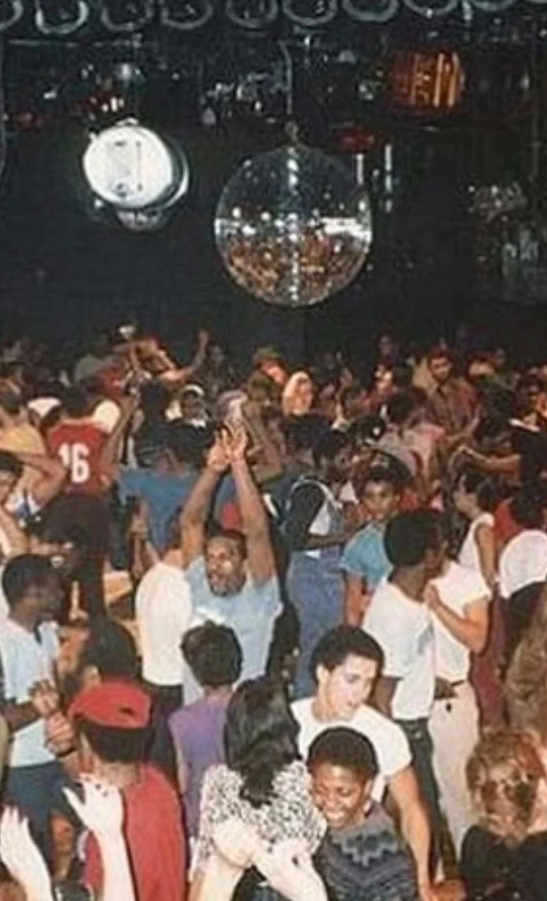 A crowded dance floor with people dancing under a large mirror disco ball, colorful outfits, and festive atmosphere, suggesting a lively party or nightclub scene from the 1970s or 1980s.