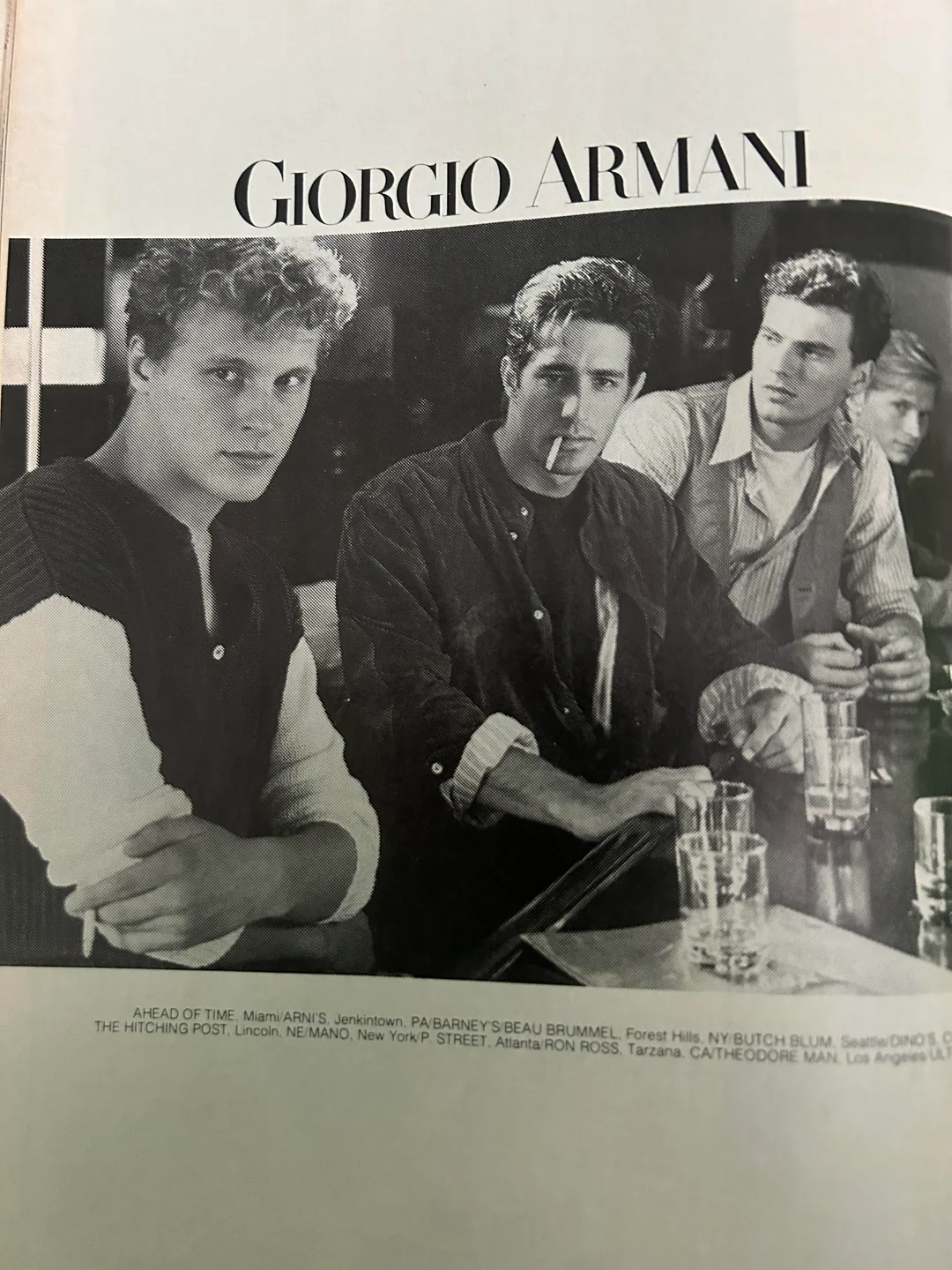 Three young men sit at a bar in a black-and-white Giorgio Armani ad. One looks at the camera, one smokes a cigarette, and the third looks away. Glasses and drinks are on the bar in front of them.