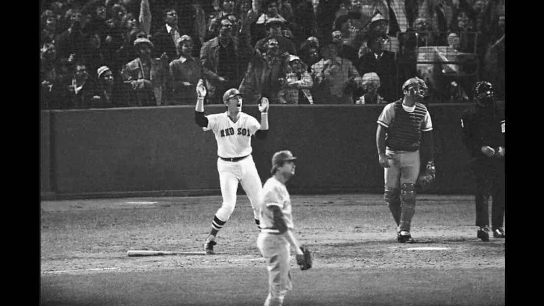 A baseball player in a Red Sox uniform stands on the field with arms raised, while a crowd watches from the stands. Other players and an umpire are visible near home plate.