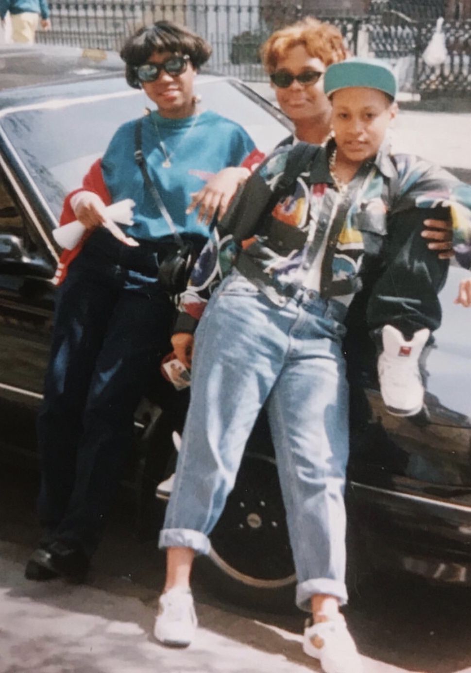Three people pose confidently by a black car. They wear retro 80s or 90s outfits, sunglasses, and sneakers. One sits on the hood, while the others lean against the car, all smiling and relaxed outdoors.