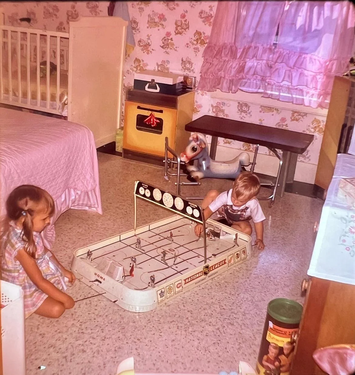 Two young children play tabletop hockey on the floor of a vintage bedroom with pink decor, toys, a crib, and a play kitchen in the background.