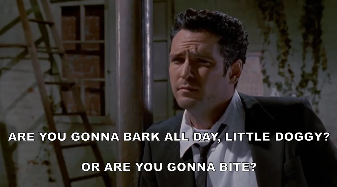 A man in a suit stands indoors, looking skeptical. White caption text reads: “ARE YOU GONNA BARK ALL DAY, LITTLE DOGGY? OR ARE YOU GONNA BITE?”