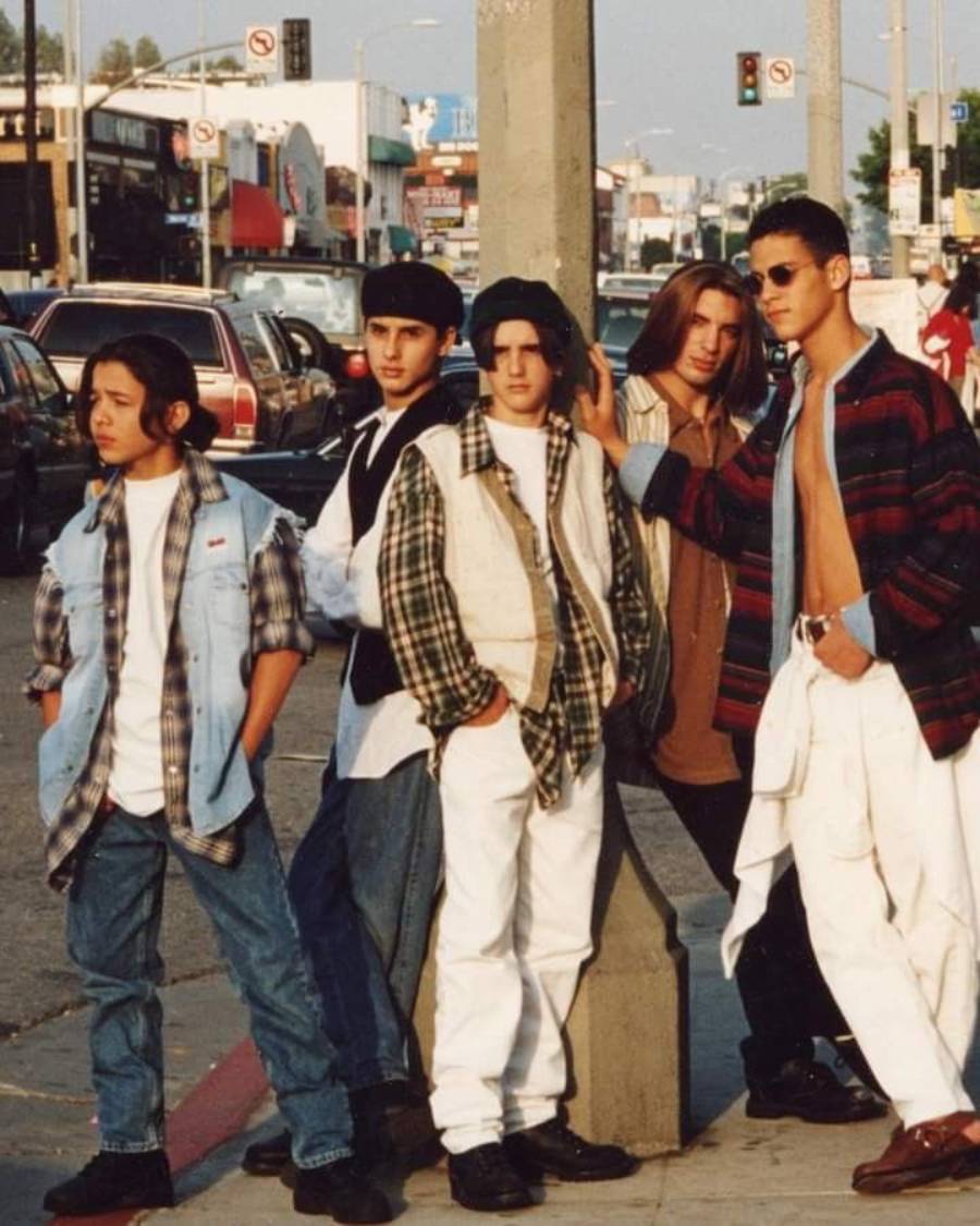 Five teens in 1990s streetwear pose casually on a city sidewalk. They wear layered shirts, baggy jeans, and sunglasses, with cars and buildings in the background.
