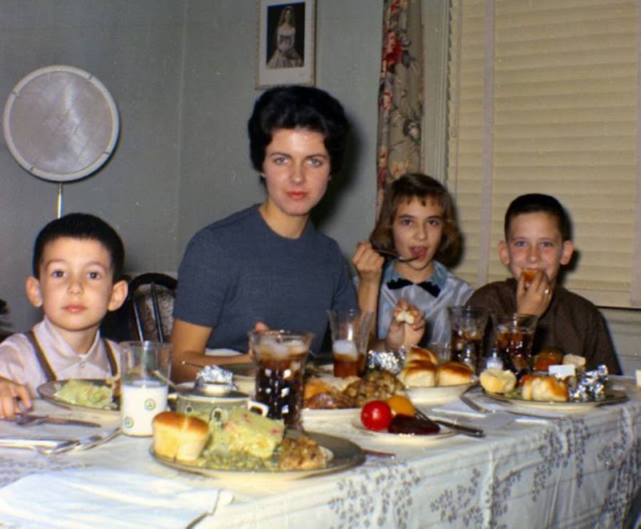 A woman and three children sit at a dining table set with food and drinks, appearing to enjoy a meal together in a cozy, warmly lit room.