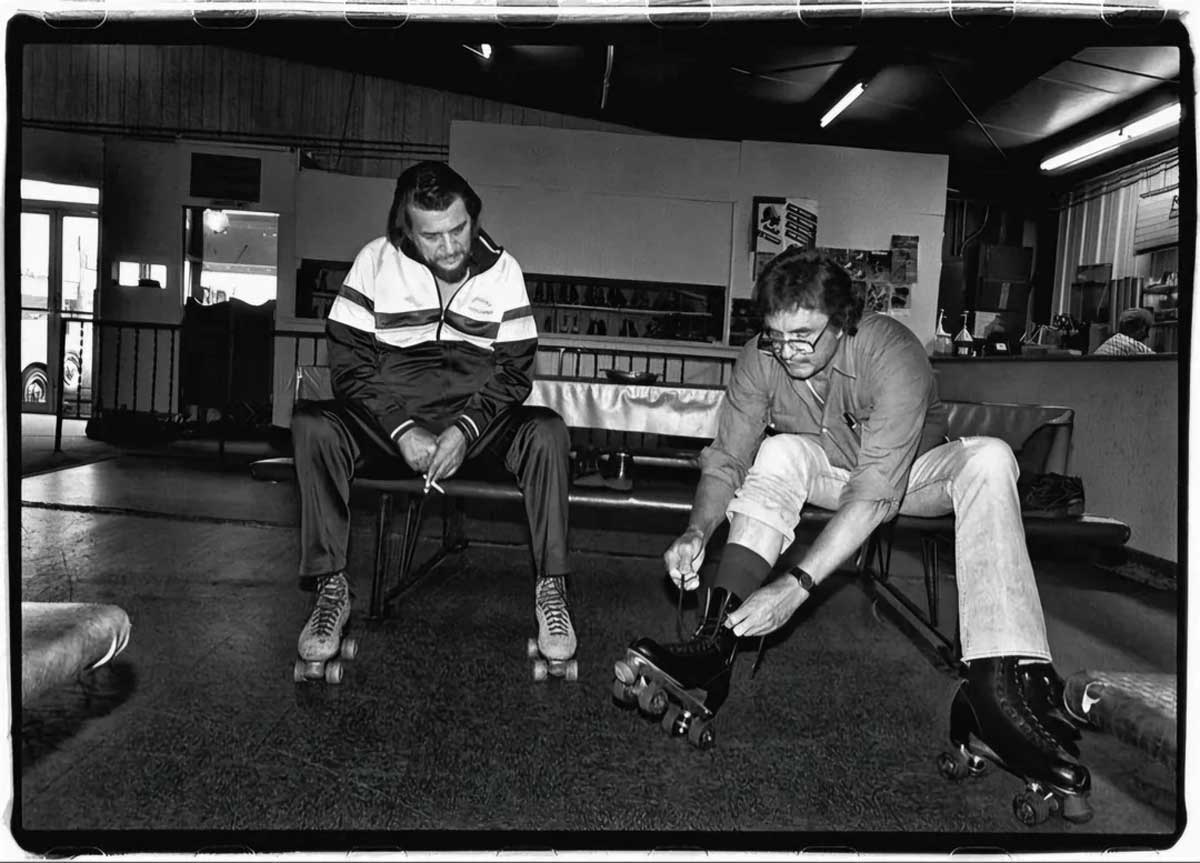Two men sit on benches in a roller rink. One man, already wearing roller skates, watches as the other man bends down to fasten his own skates. The room has benches, shelves, and various skating equipment.