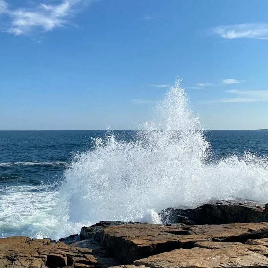 Waves crash against rocky shore under a clear blue sky, sending a tall spray of white water into the air.