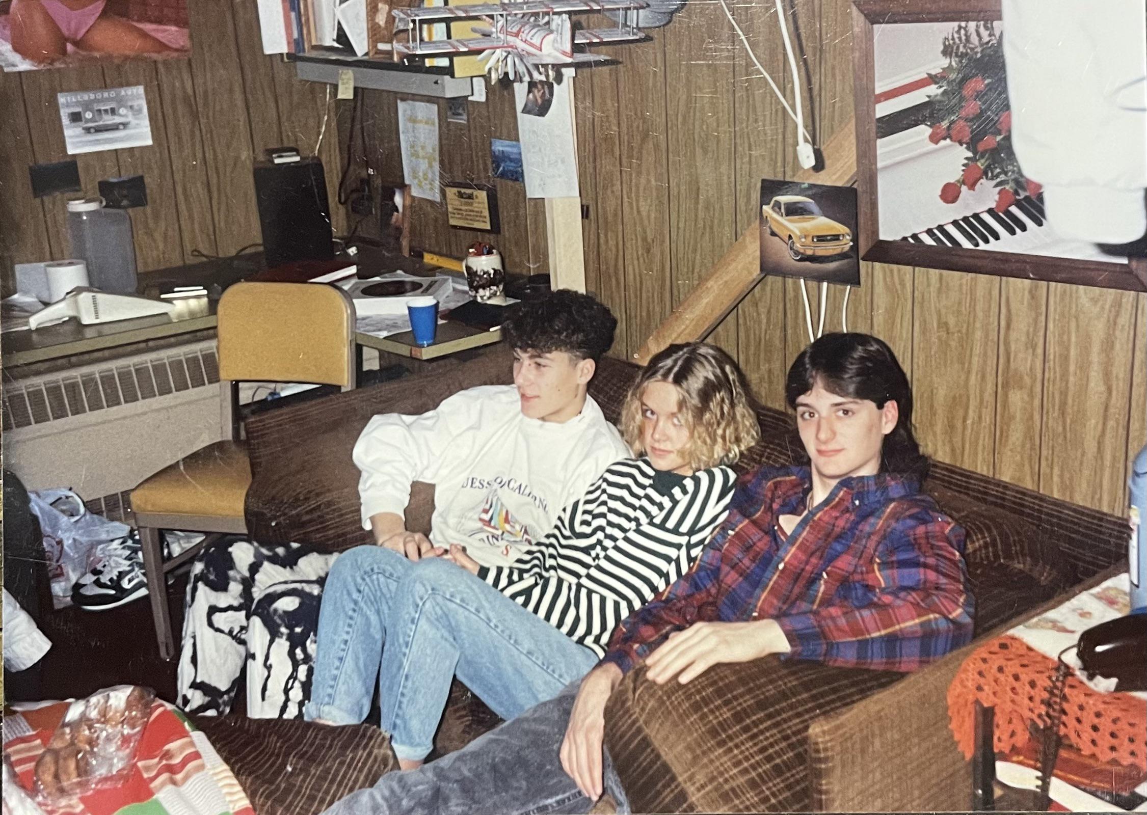 Three teenagers sit close together on a brown couch in a wood-paneled room decorated with posters, papers, and a yellow chair. The atmosphere appears casual and relaxed.