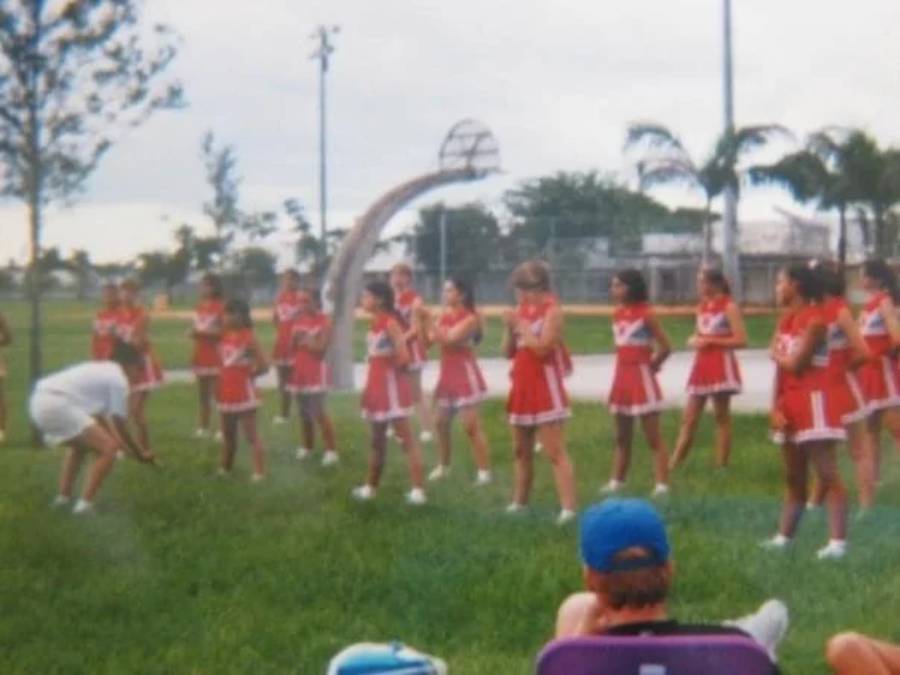 A group of cheerleaders in red uniforms performs outdoors on grass. A coach bends over in front of them. A person in a blue cap sits in a chair in the foreground. Palm trees and a basketball hoop are in the background.