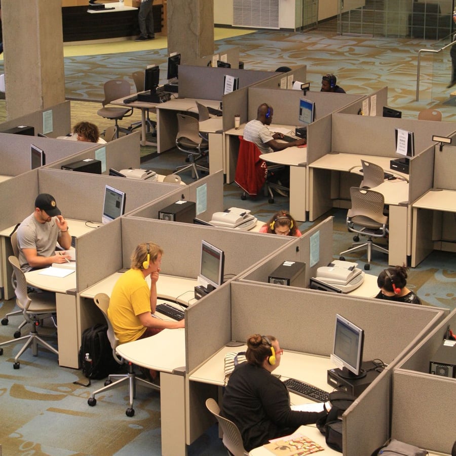 People sit individually in cubicles at computers in a modern library or office space, working quietly. The cubicles are arranged in rows, and most users wear headphones. The environment looks focused and studious.