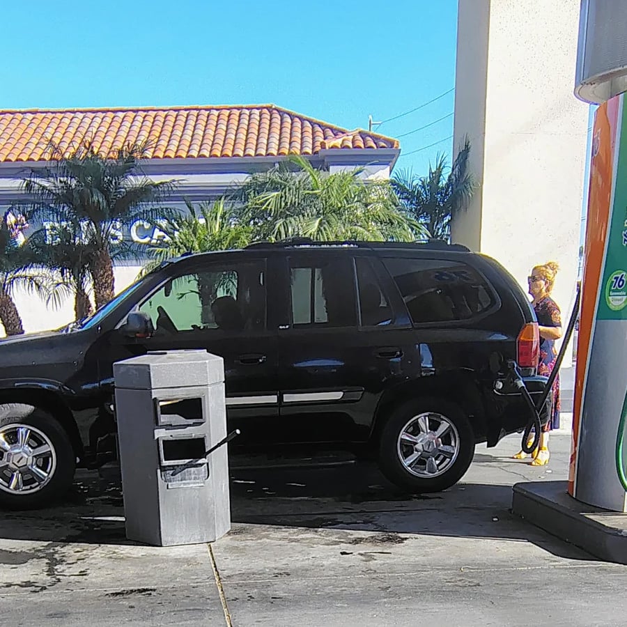 A black SUV parked at a gas station pump with a person in bright clothing standing nearby. Palm trees and a tile-roofed building are visible in the background under a clear blue sky.