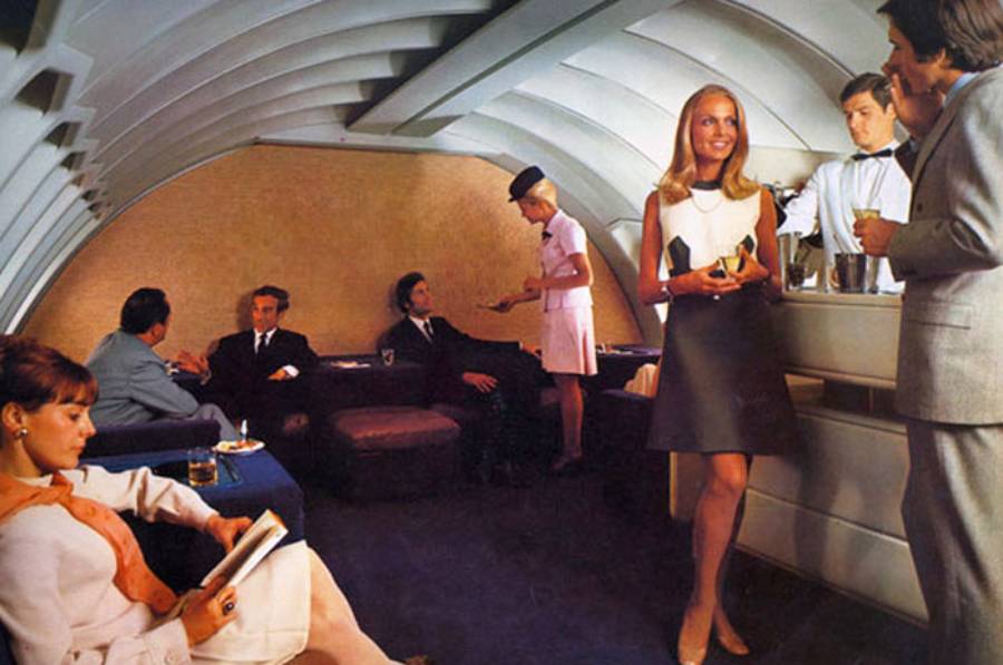 People relax and socialize in a stylish, retro airplane lounge with curved walls. A flight attendant serves guests, while others talk, read, or enjoy drinks. The setting appears elegant and sophisticated.
