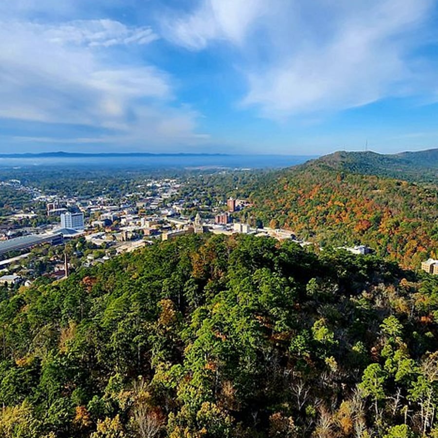 Aerial view of a city surrounded by forested hills with scattered autumn foliage under a blue sky with wispy clouds.